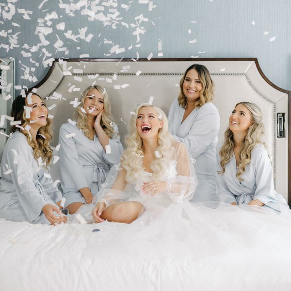 Bride and her bridesmaids sitting on a bed throwing confetti