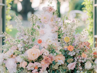 3 Tier White Wedding Cake Decorated and Surrounded by Light Pink, Orange, and Yellow Flowers