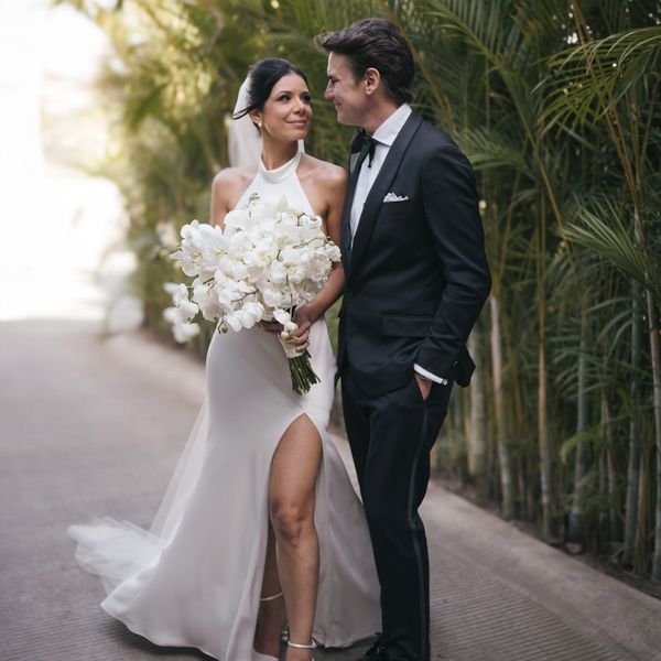 bride wearing a halter-neck wedding dress while holding a bouquet of white flowers; groom wearing a black tux