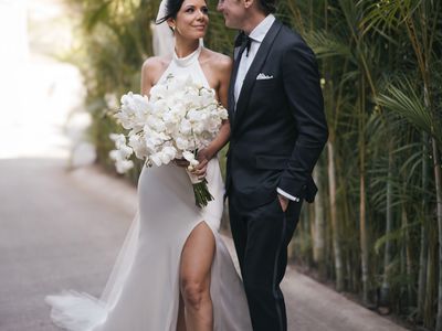 bride wearing a halter-neck wedding dress while holding a bouquet of white flowers; groom wearing a black tux