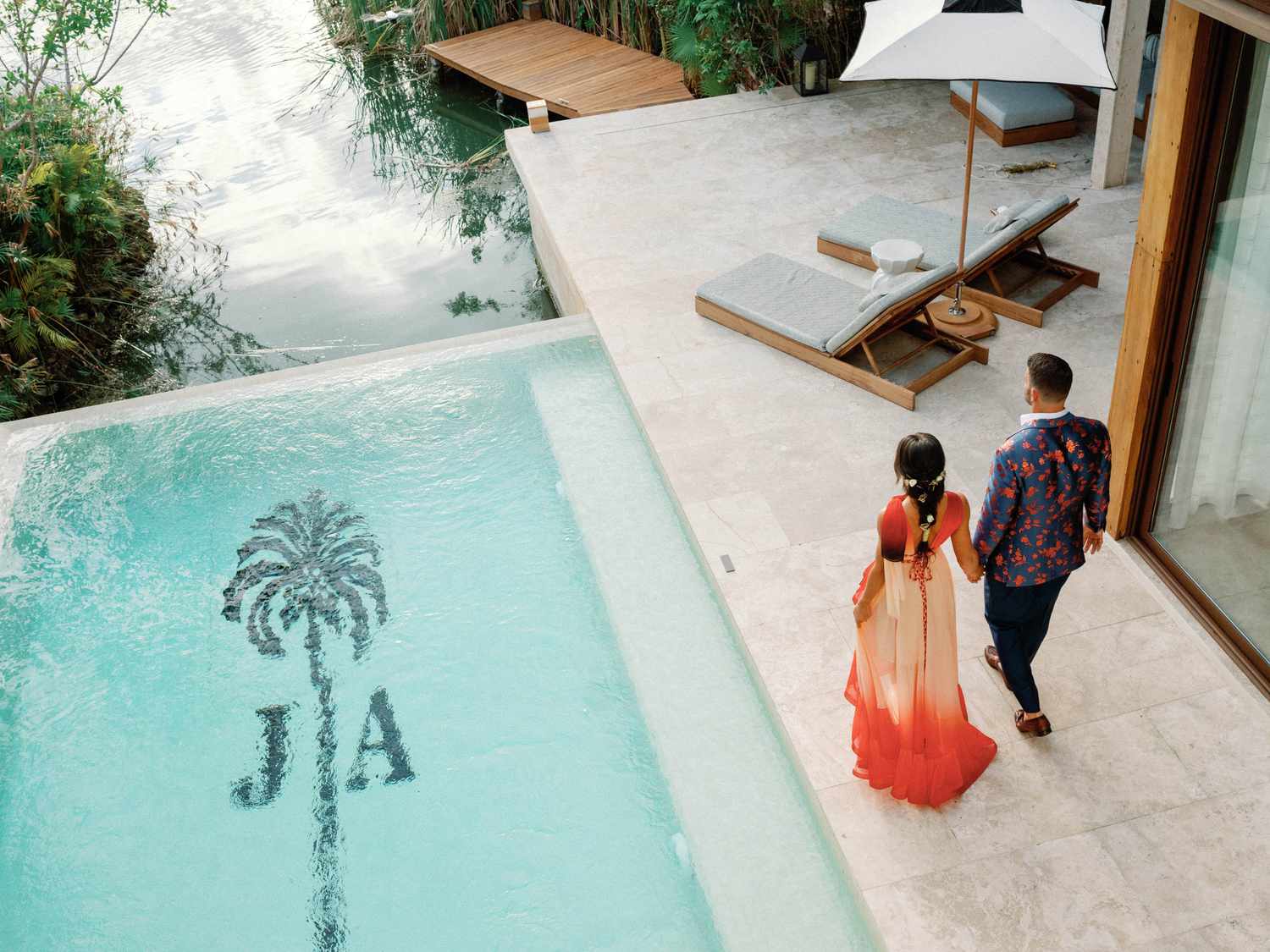 bride and groom in colorful attire walking next to pool with monogram