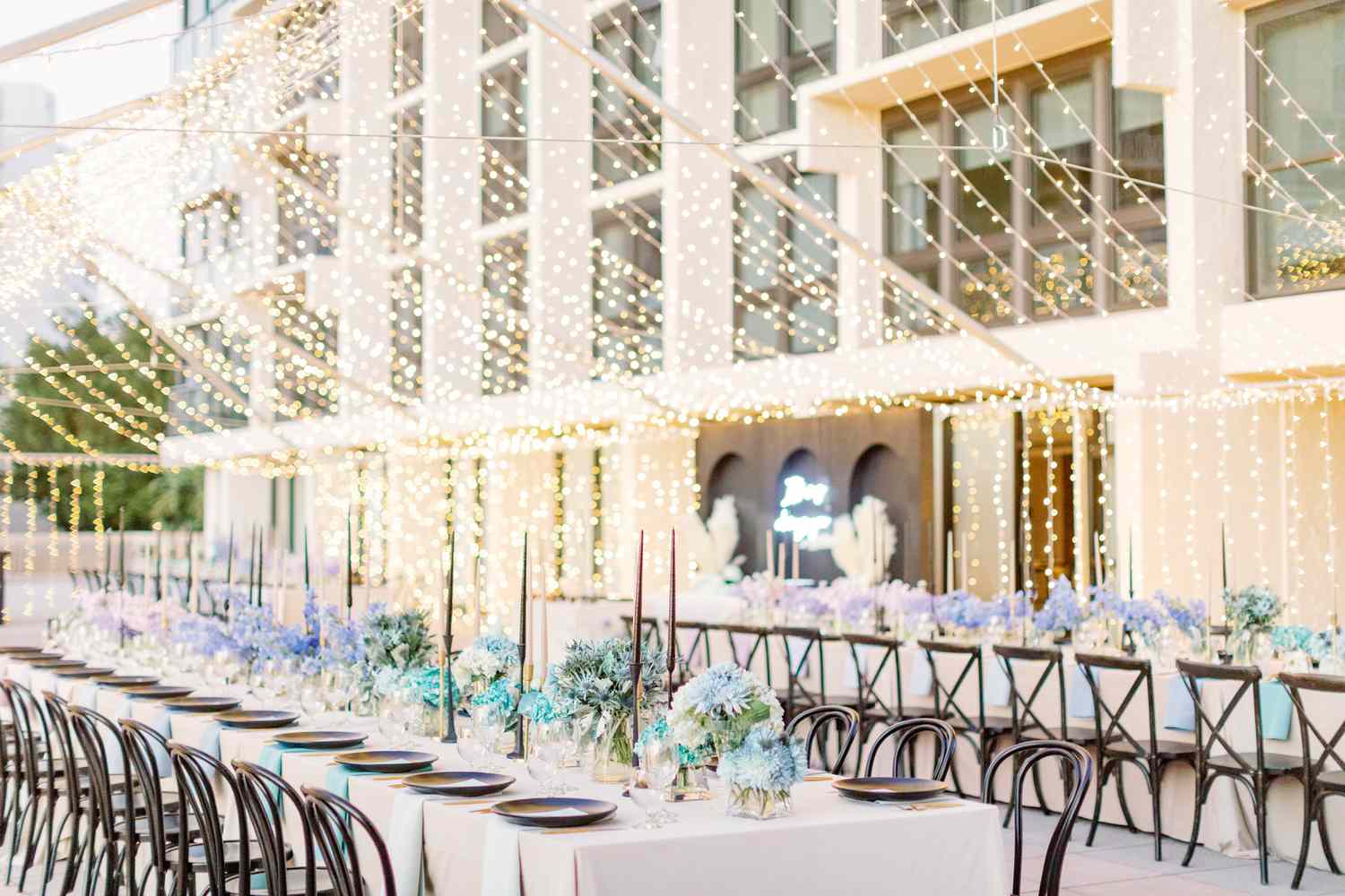Purple Flower Decor Next to Purple Table Setting on White Tables Under String Lights at Wedding Reception