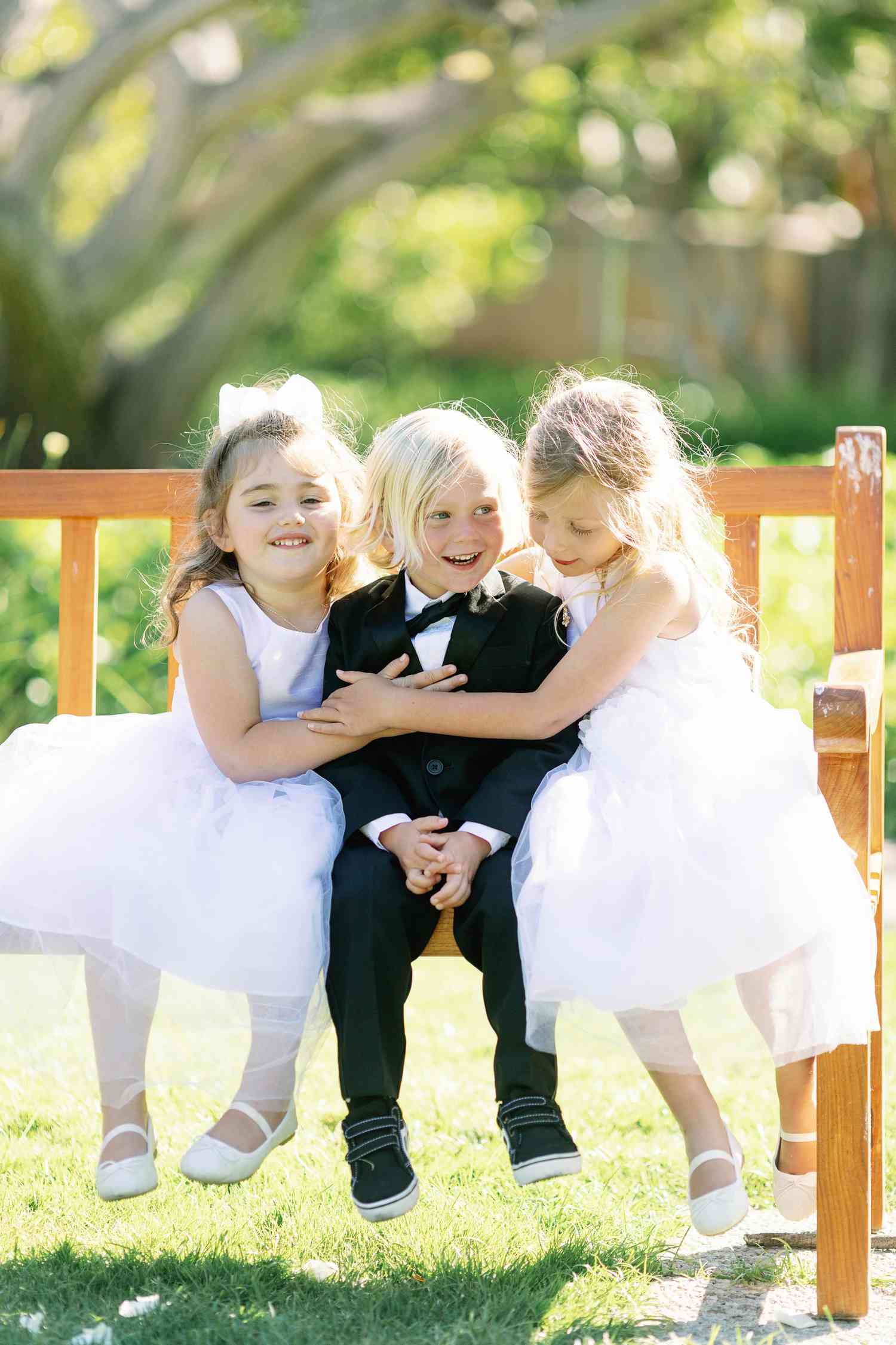 Ring Bearer in Black Tuxedo and Flower Girls in White Dresses Hugging on a Bench