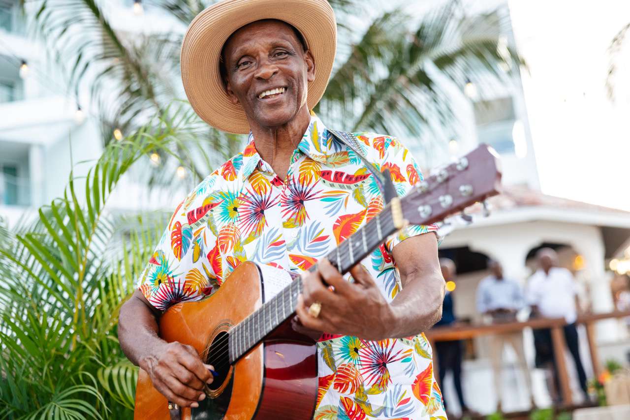Close Up of Man in Colorful Tropical Button Up and Hat Playing the Guitar at Wedding Cocktail Hour