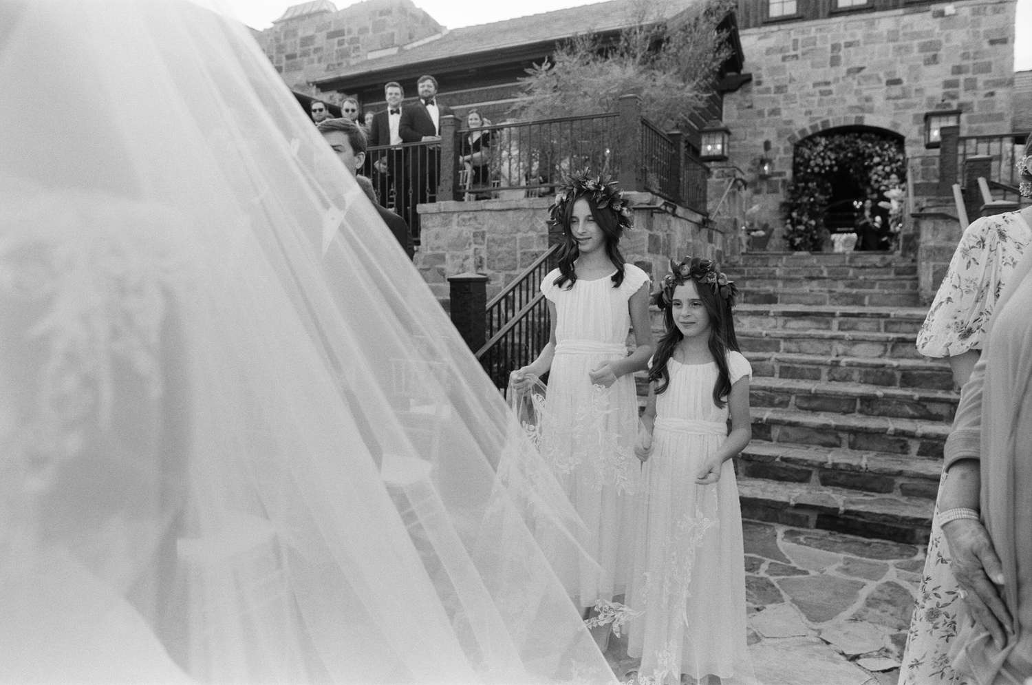 Young Girls in White Dresses and Flower Crowns Holding Brides Veil Down Aisle