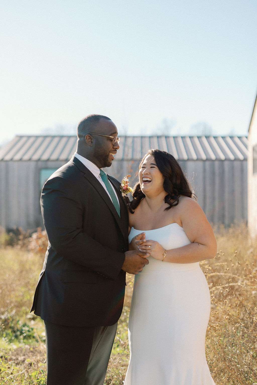Bride in Strapless Wedding Dress Posing With Groom in Black Suit in Grassy Field