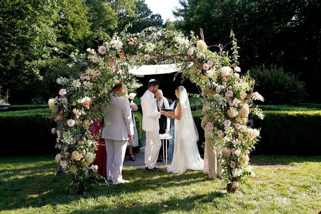 a bride and groom under a chuppah covered by a floral arch of pink roses