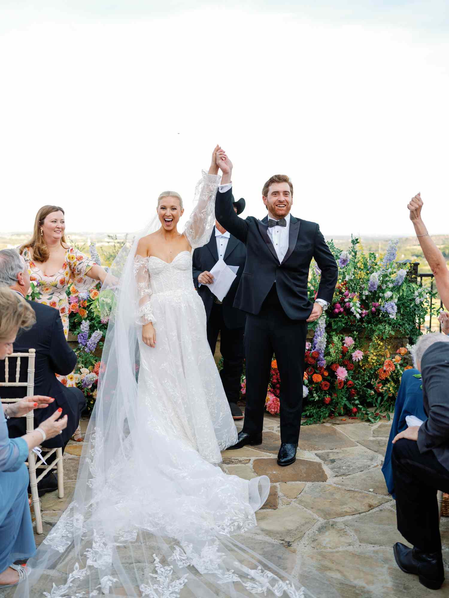 Bride in White Lace Wedding Dress With Long Veil Holding Hands with Groom in Black Tuxedo
