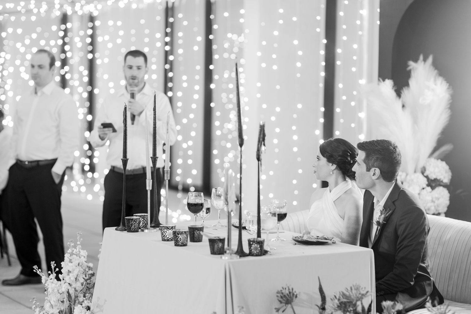 Black and White Portrait of Bride in Halter Top Wedding Dress At Main Table With Groom in Suit While Man Gives Speech