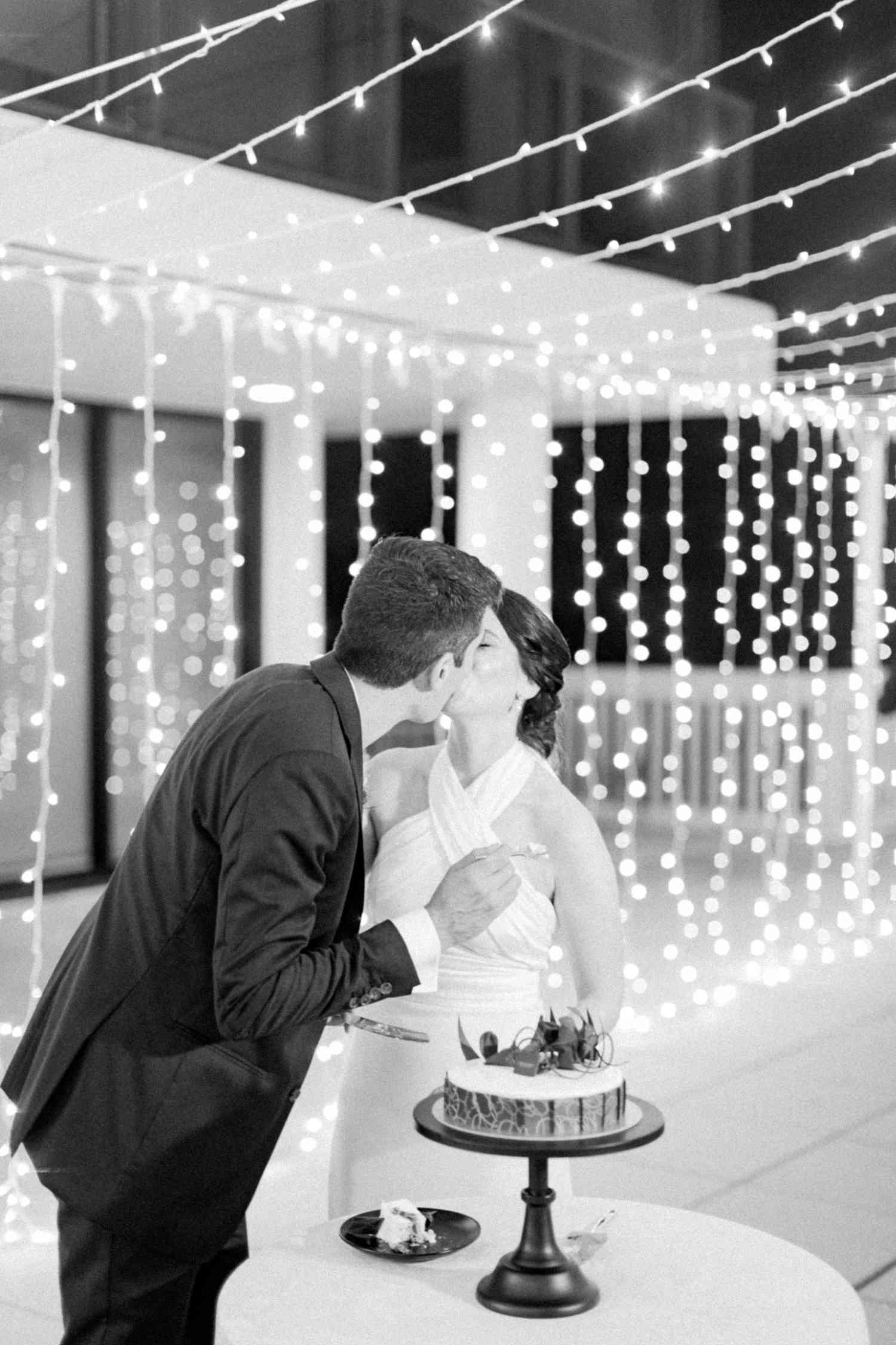 Black and White Portrait of Bride in Halter Top Wedding Dress Kissing Groom in Suit by Cake