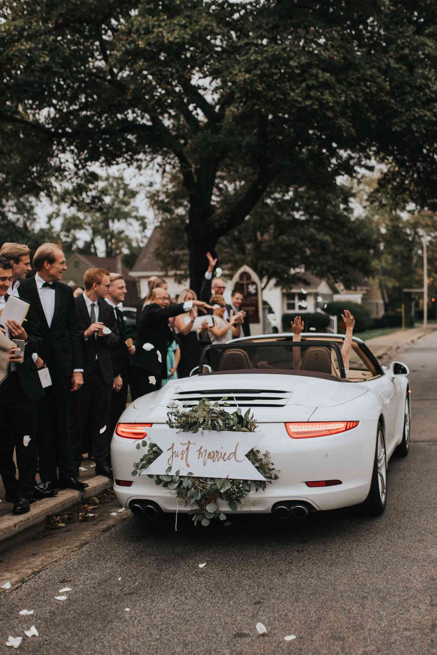 Bride and groom riding in a convertible decorated with a wreath of eucalyptus while guests cheer