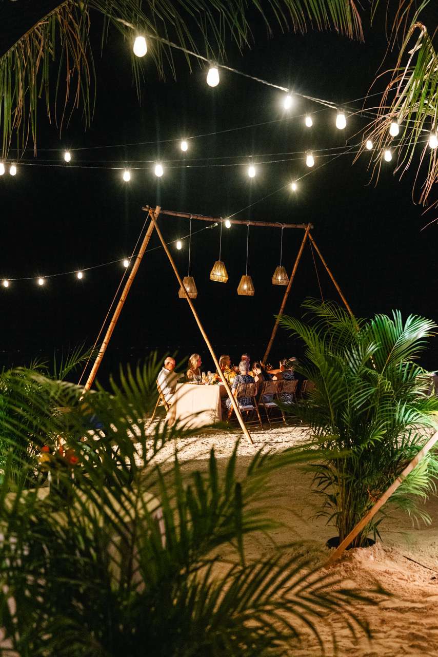 Reception Guests Seated at Small Reception Table on Beach With Overhead String Lights and Tropical Plants