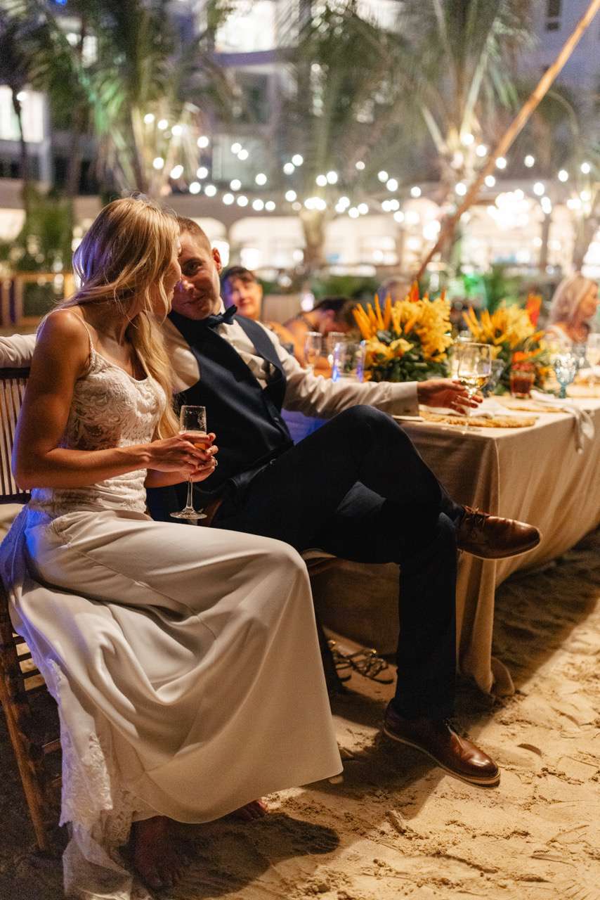 Bride in Wedding Dress Seated Next to Groom in Tuxedo Holding Champagne at Reception Table