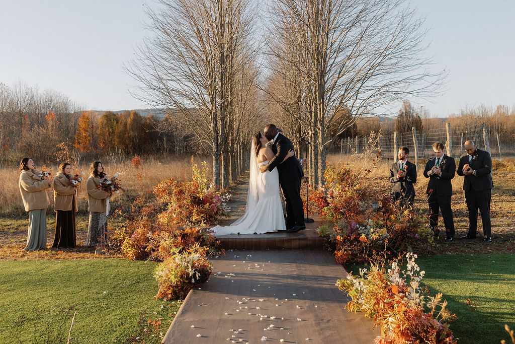 Bride in Strapless Dress with Veil Kissing Groom in Black Suit at Outdoor Altar