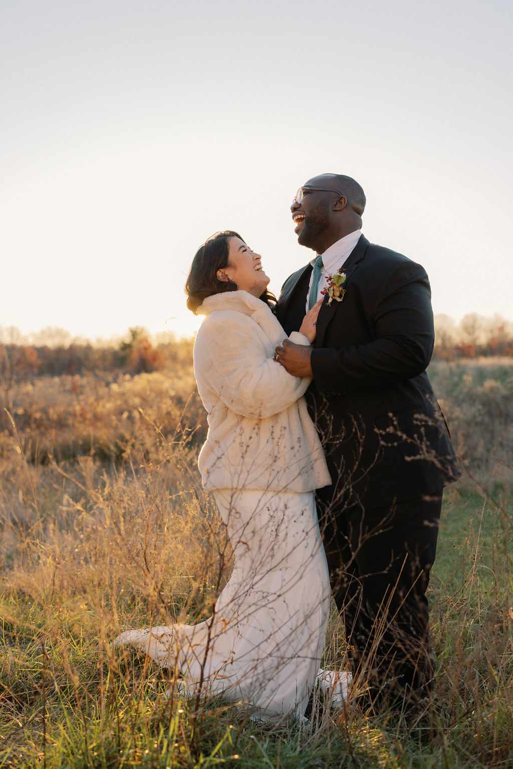 Bride in White Dress and White Coat Laughing With Groom in Black Suit in Grassy Field
