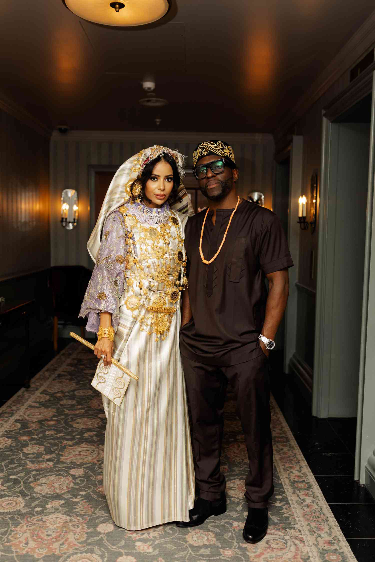 Bride in Traditional White and Gold Dress With Gold Jewelry and Veil Posing With Groom in Brown Outfit With Gold Cap And Gold Chain in Dimly Lit Room