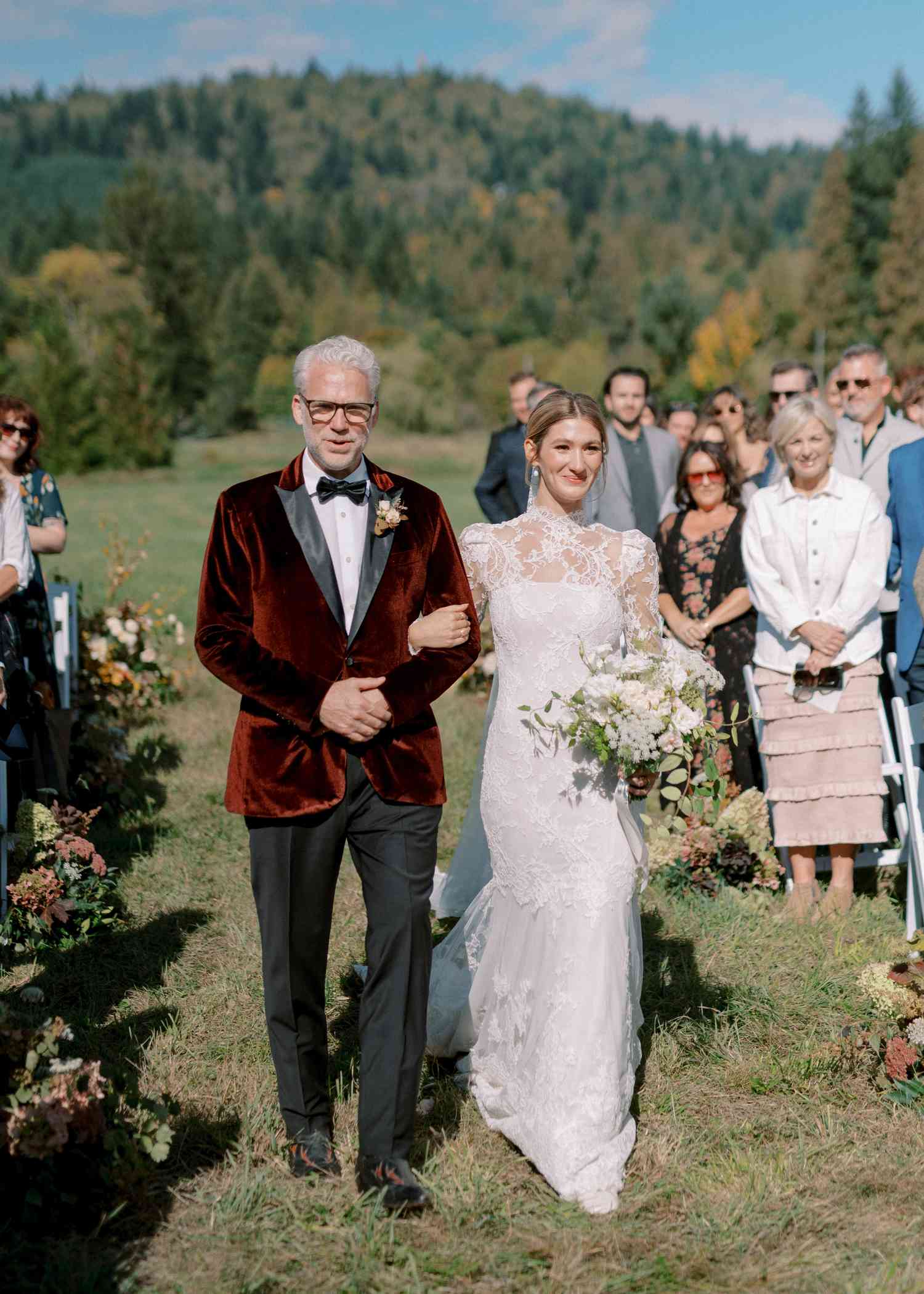 Bride in Lace Wedding Dress Holding White Bouquet Walking Down Aisle With Father in Red Velvet Tuxedo