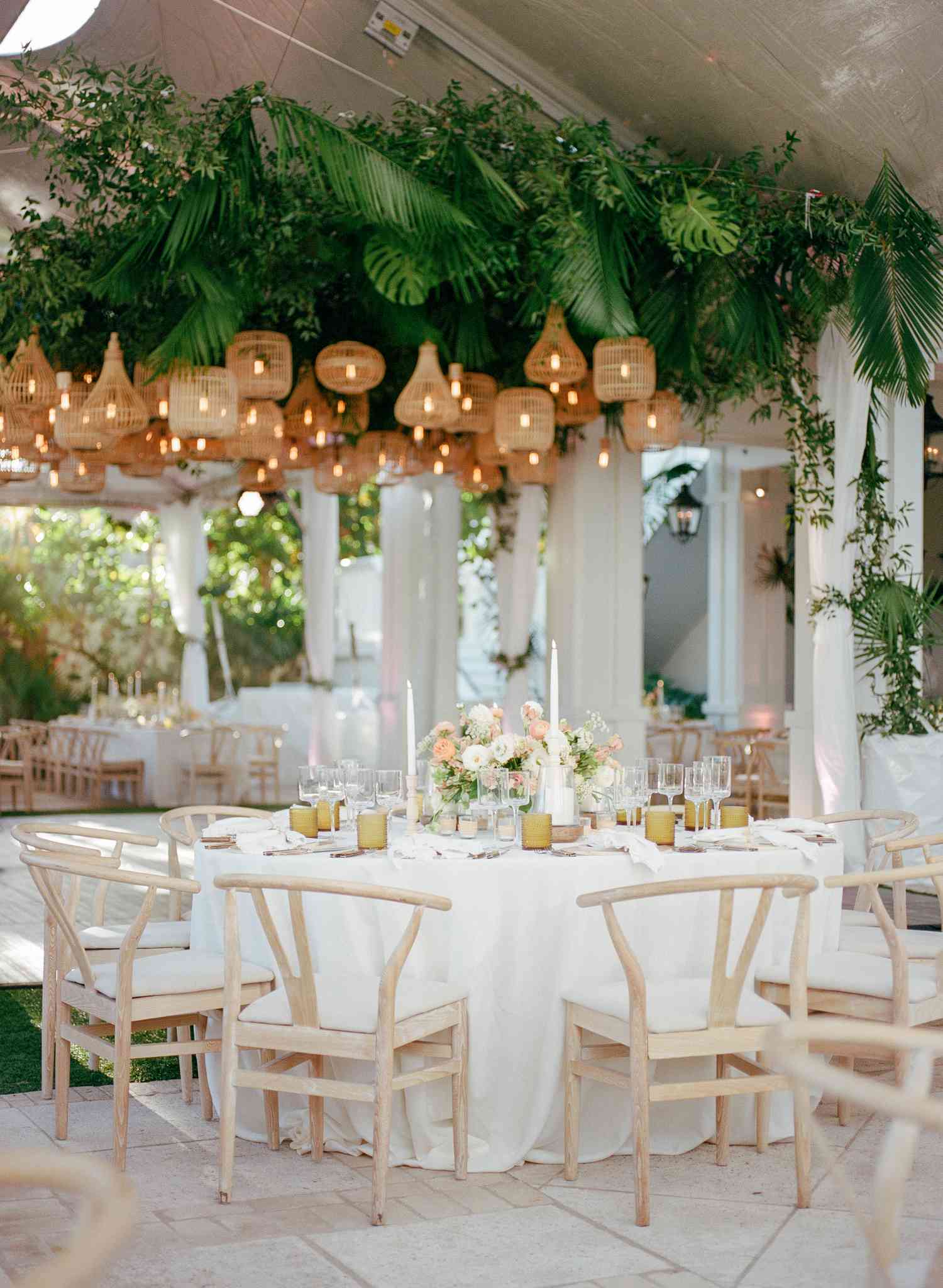 Round Wedding Reception Table With White Tablecloth and Washed Wood Chairs Positioned Under Woven Lanterns and Tropical Greenery Installation