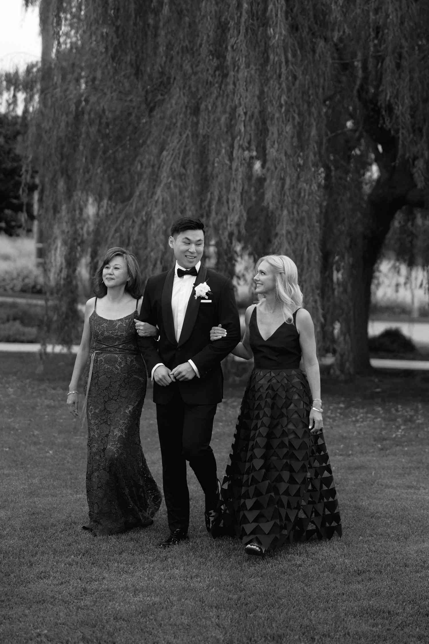 Black and White Image of Groom in Tuxedo Locking Arms With Women in Dresses in Front of Willow Tree