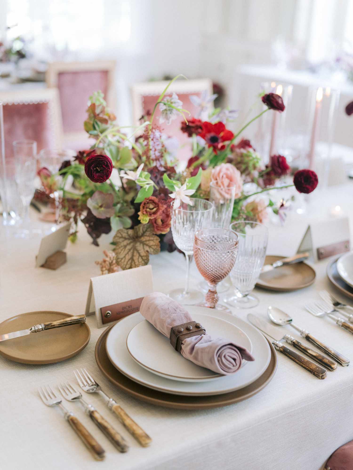Floral centerpieces of anemones, ranunculus, roses, and greenery paired with leather accents at place settings with bone flatware, colored glass goblets and velvet details