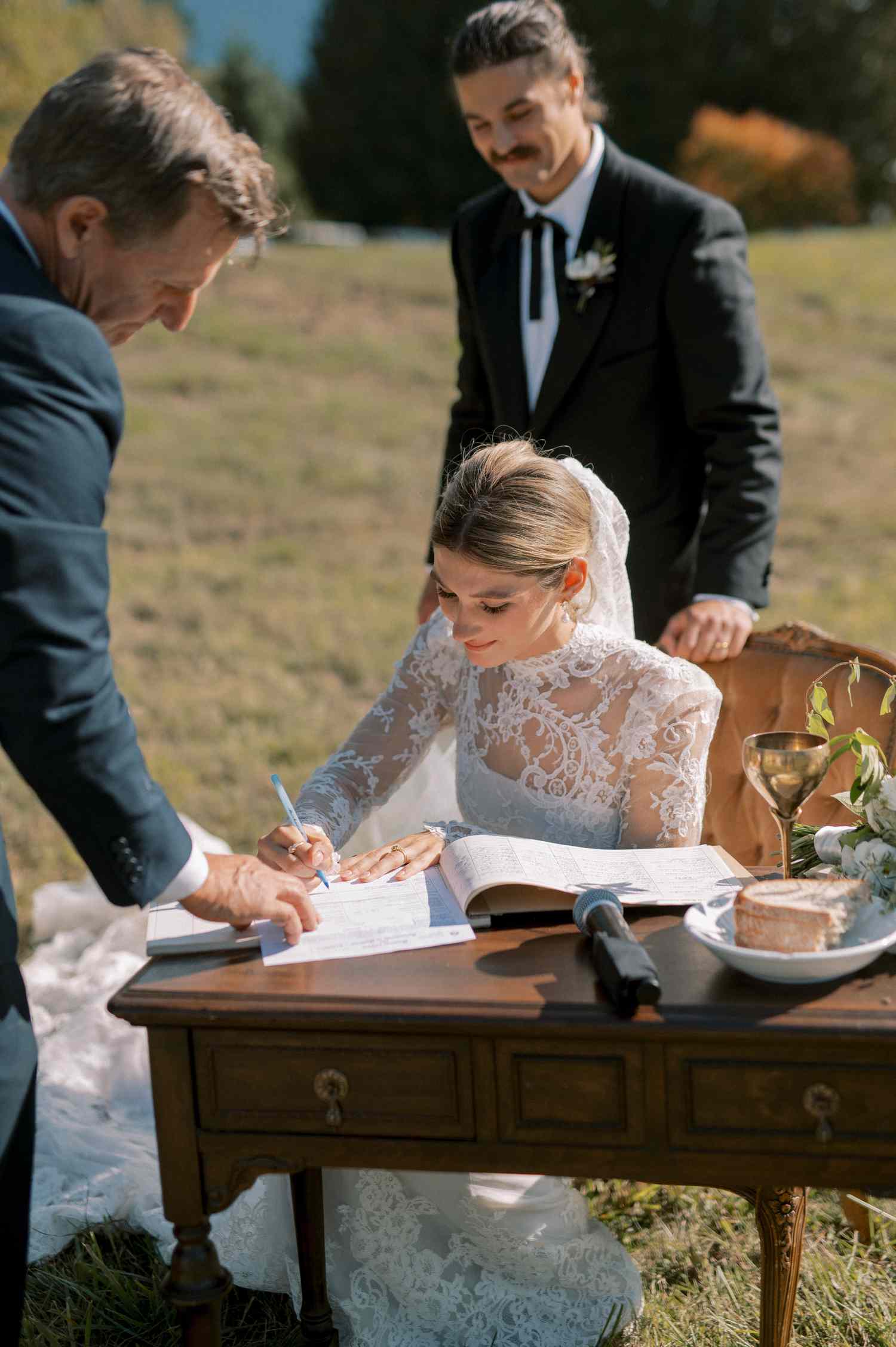 Bride in Lace Wedding Dress Signing Marriage License With Groom in Black Tuxedo Standing Behind Her