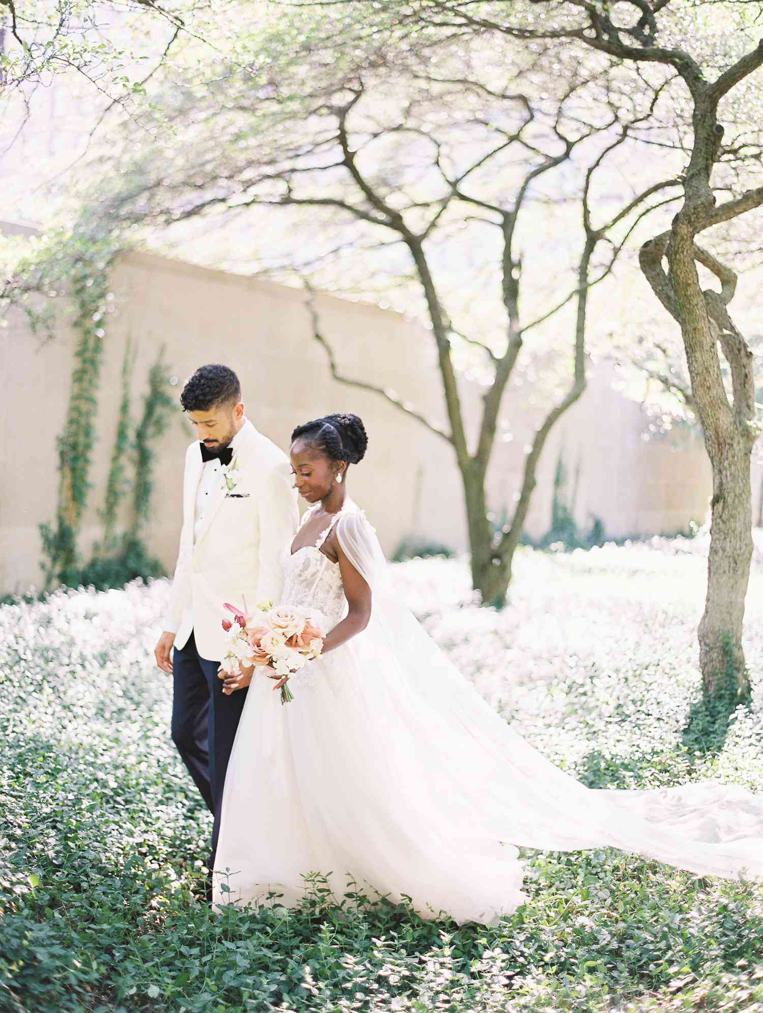 Groom in White Tuxedo and Bride in Lace Wedding Dress Holding Hands and Walking in Floral Field