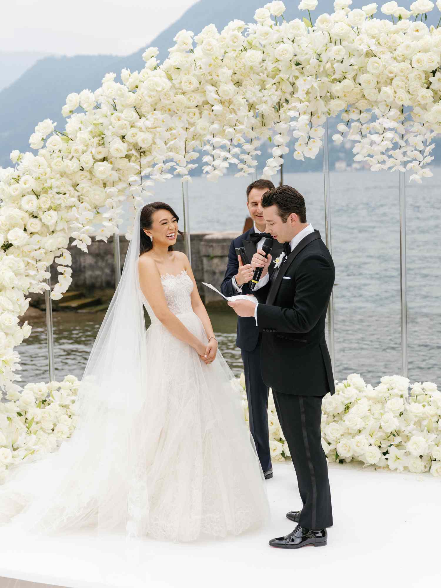 Groom in Black Tuxedo Reading Vows to Bride in White Wedding Dress and Veil at White Flower Altar By Ocean