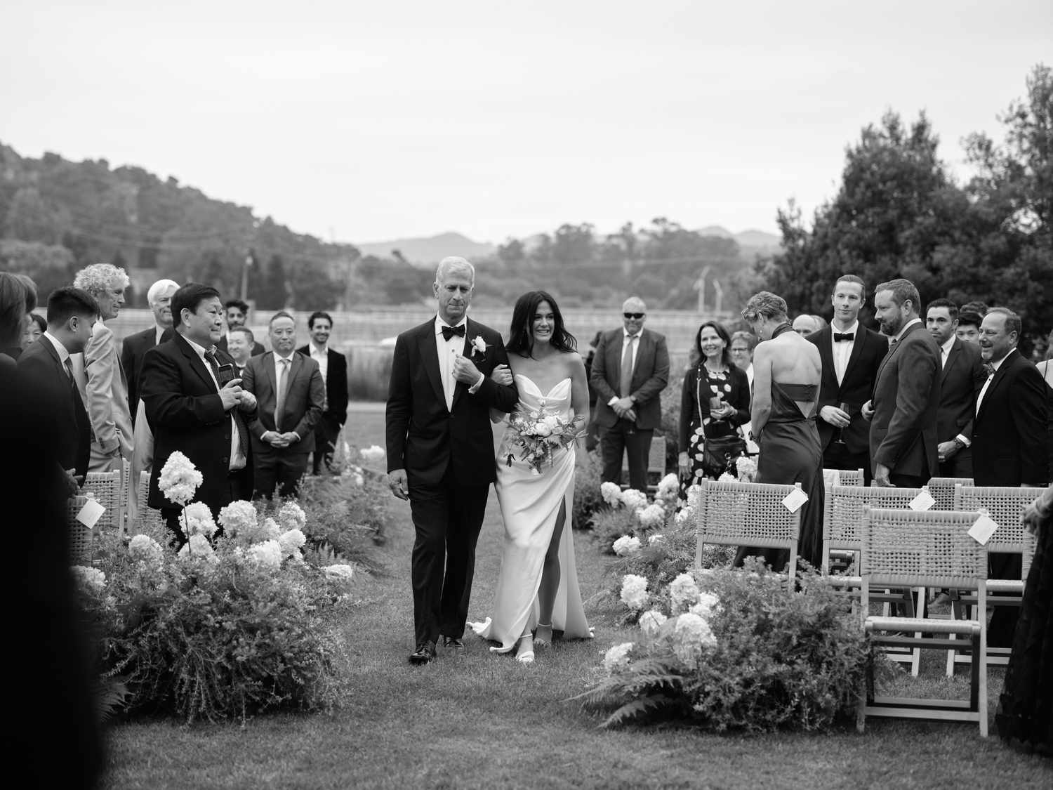 Black and White Portrait of Bride in Wedding Dress Walking Down Aisle with Father in Tuxedo