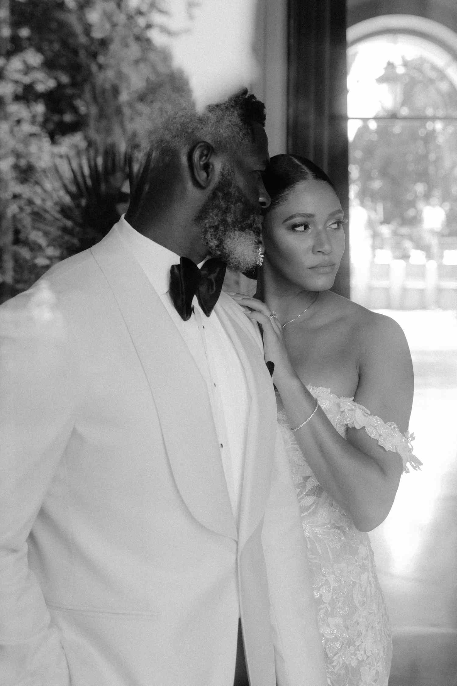Black and White Image of Bride in Lace Wedding Dress With Hair in Bun Posing With Groom in Tuxedo Through Window