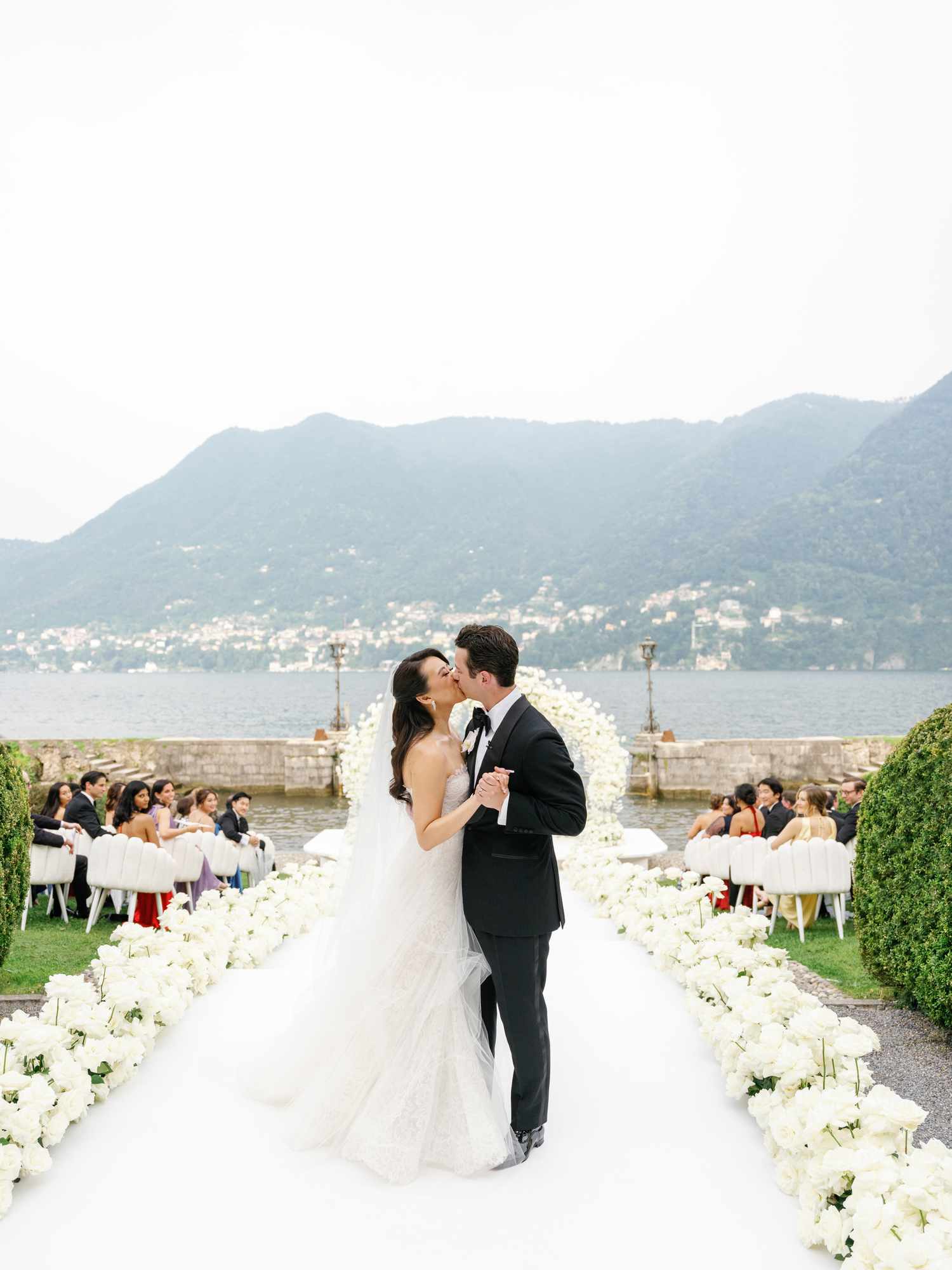 Bride in White Wedding Dress and Veil Kissing Groom in Black Tuxedo in Waterfront Aisle