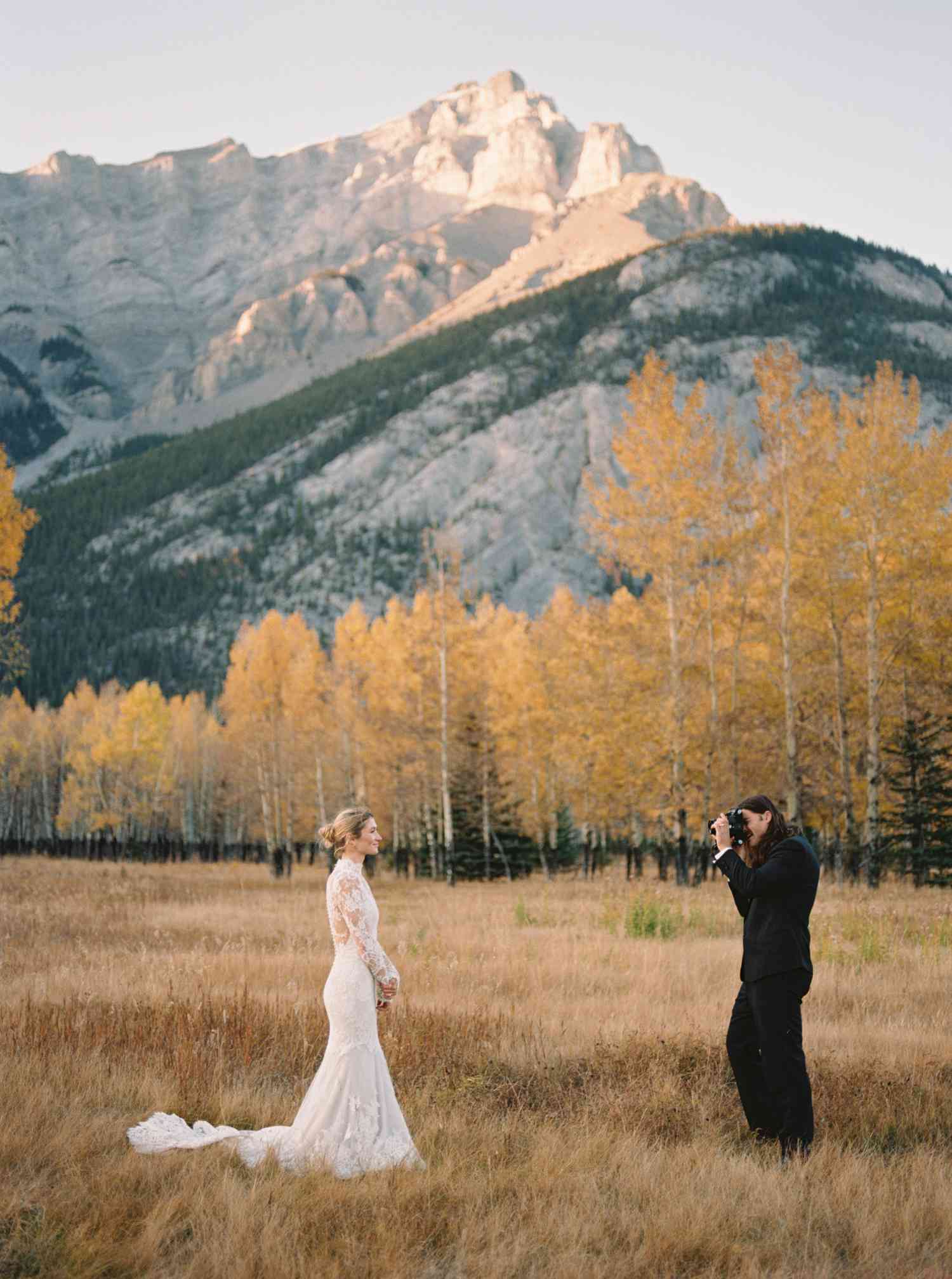 Groom in Tuxedo Taking Portrait of Bride in Lace Wedding Dress in Grass Field With Mountains in Background