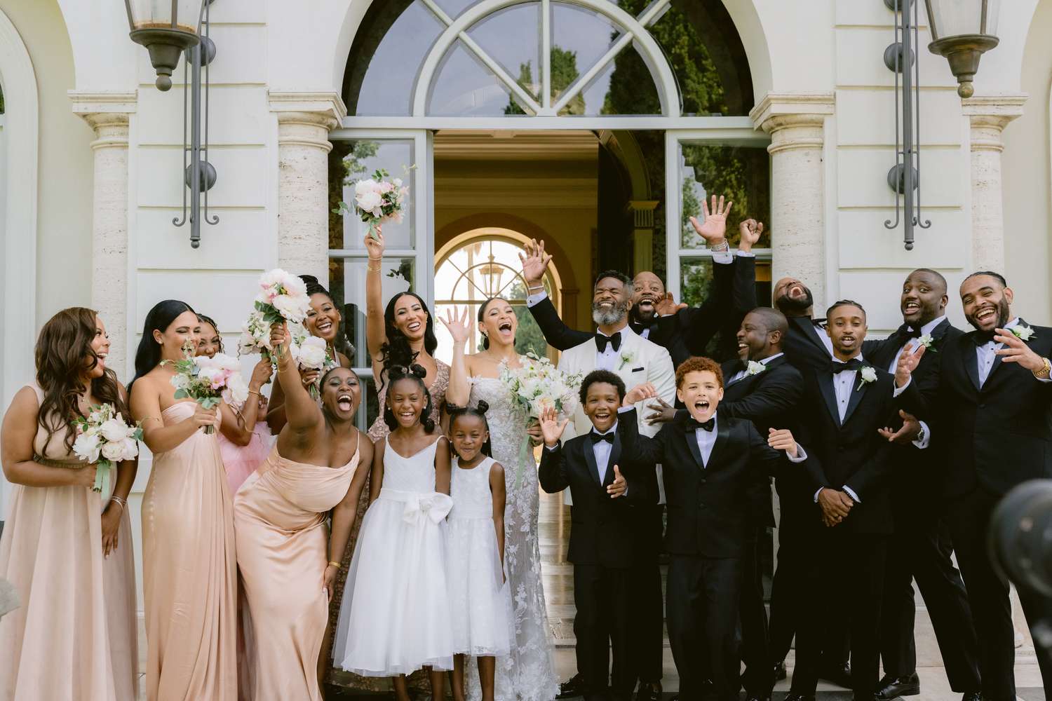 Bride in Lace Wedding Dress and Groom in Black and White Tuxedo Cheering with Wedding Party in Pink Dresses and Black Tuxedos