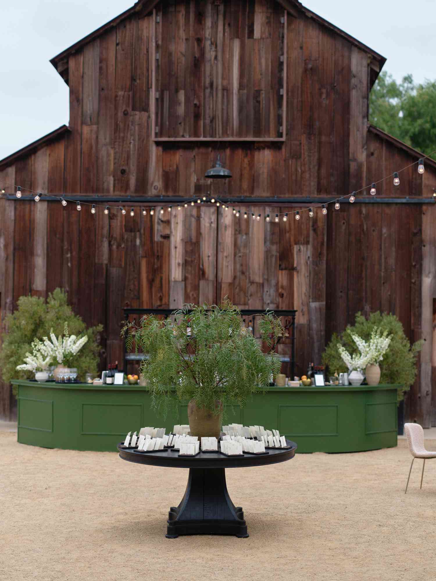 Reception Seating Chart Arranged on Black Table With Plant Decor In Front of Green Cocktail Bar by Wooden Barn