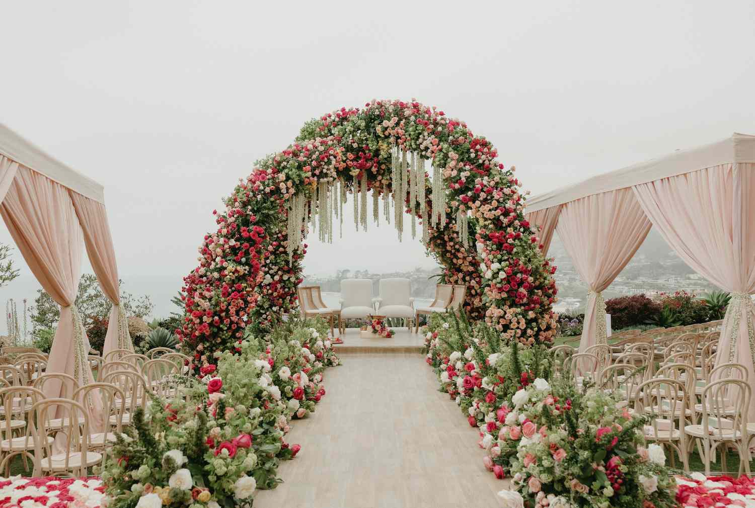 Indian Wedding Ceremony Decor With Pink and White Flowers Running Down Aisle, Pink and White Floral Arches, and Wooden Chairs Under Pink Tents with Curtains