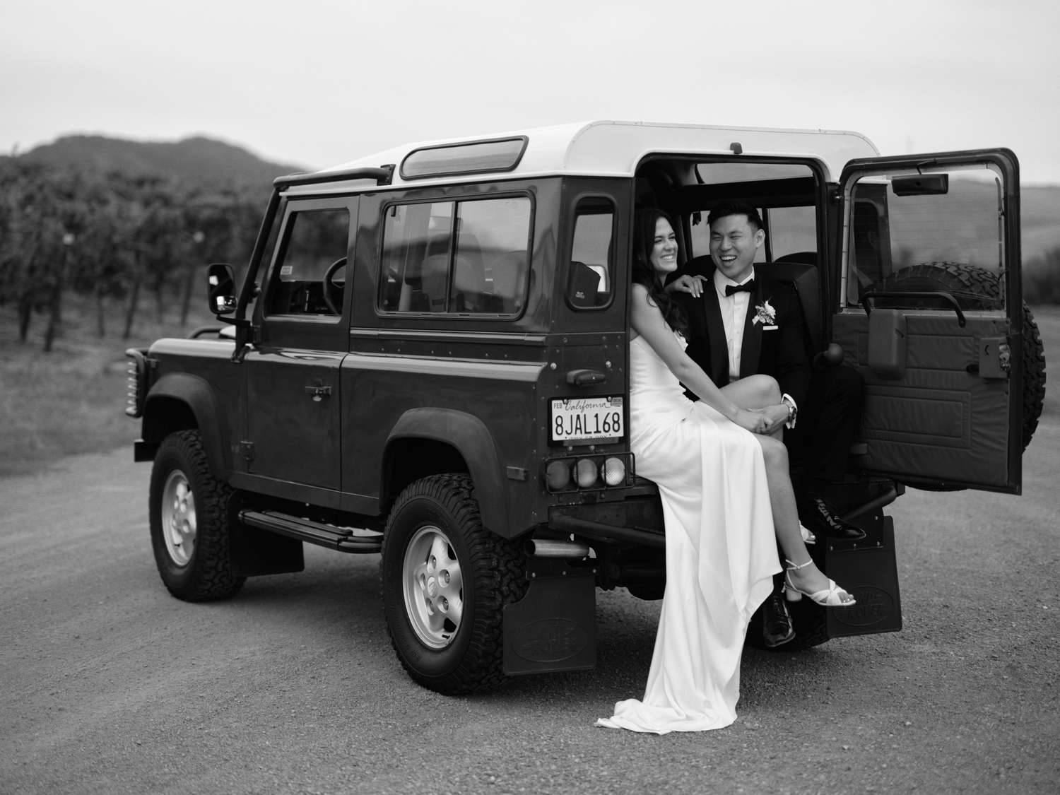 Black and White Portrait of Bride in Wedding Dress and Groom in Tuxedo in Trunk of Jeep