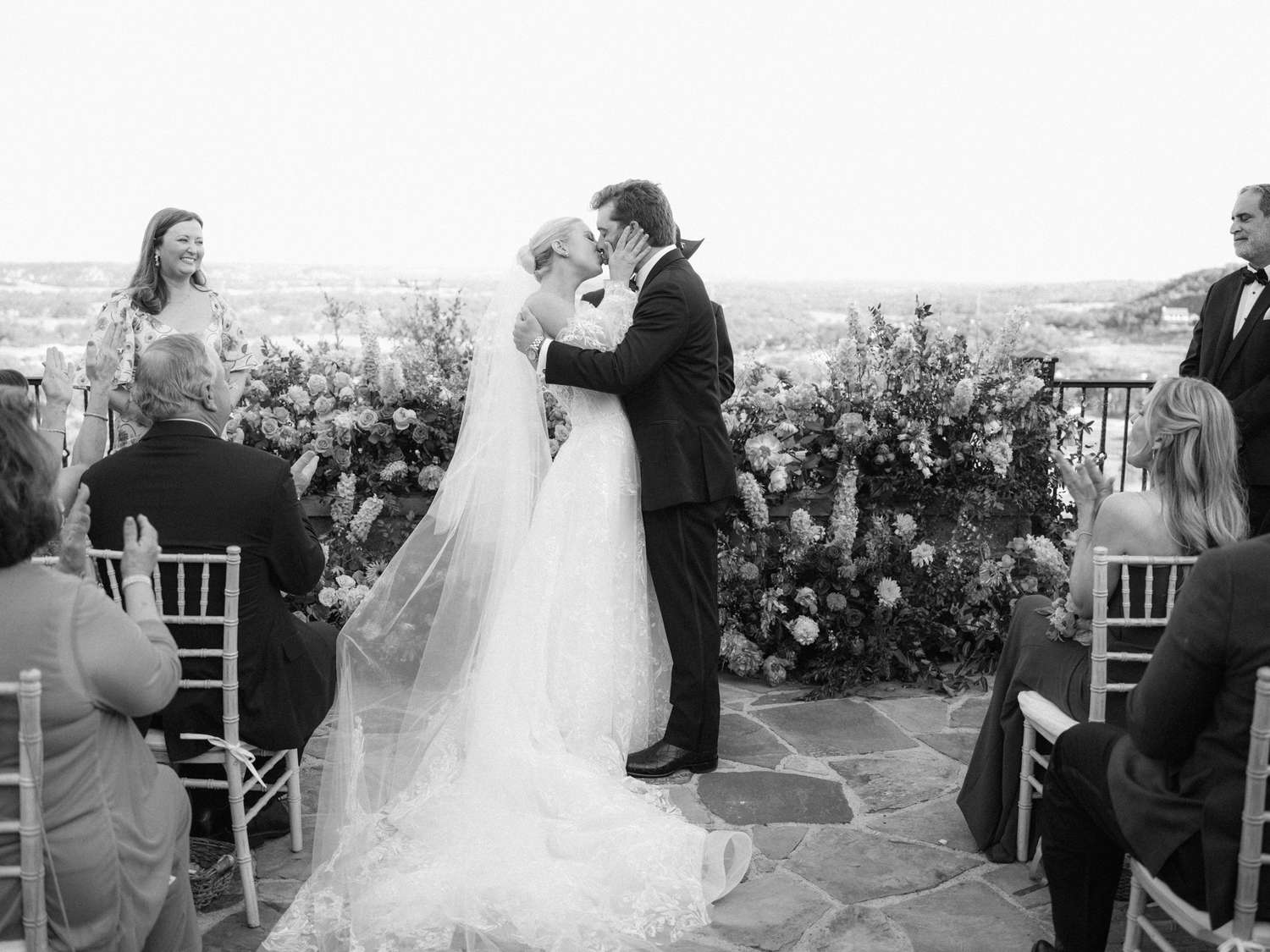 Black and White Portrait of Bride in Lace Wedding Dress and Long Veil Kissing Groom in Tuxedo at Altar