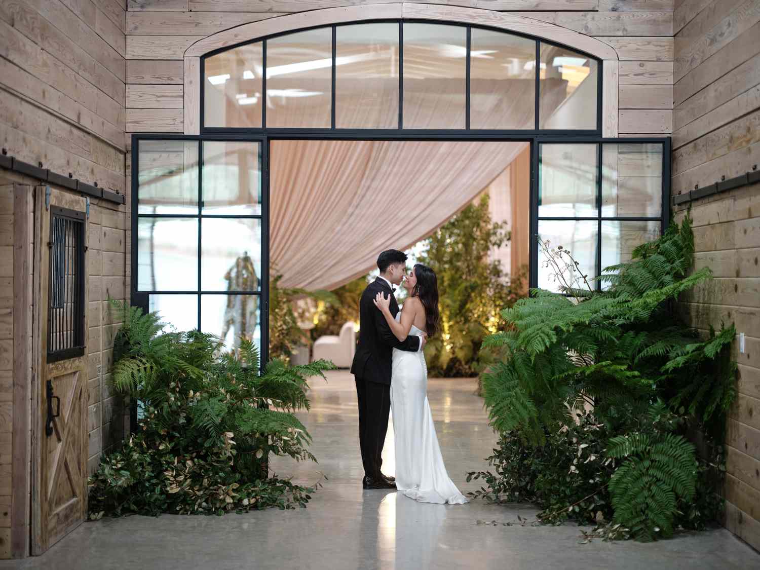 Bride in Wedding Dress Posing With Groom in Tuxedo in Front of Glass Doorway With Plant Decor