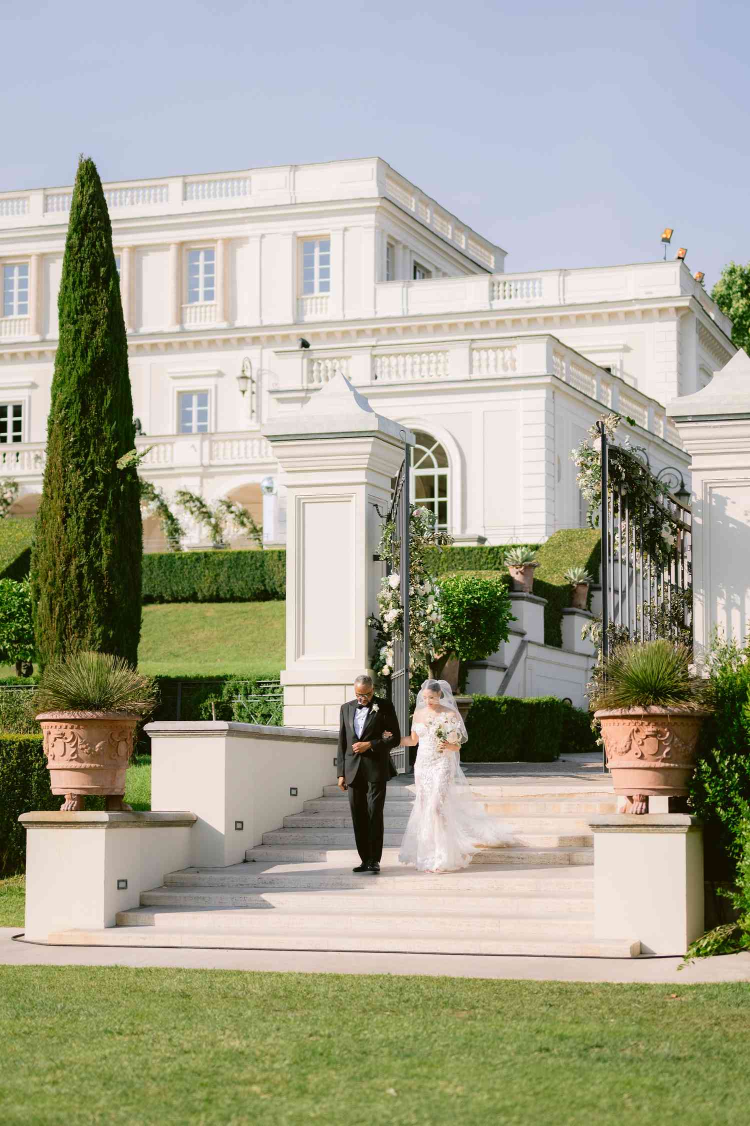Bride in Lace Wedding Dress Holding Bouquet Walking Down Steps of White Villa With Father in Black Tuxedo