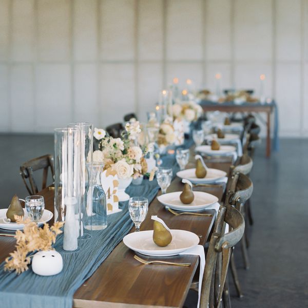 A rehearsal dinner table set with a blue runner, tall candles, white roses, and pear-shaped candle guest favors.