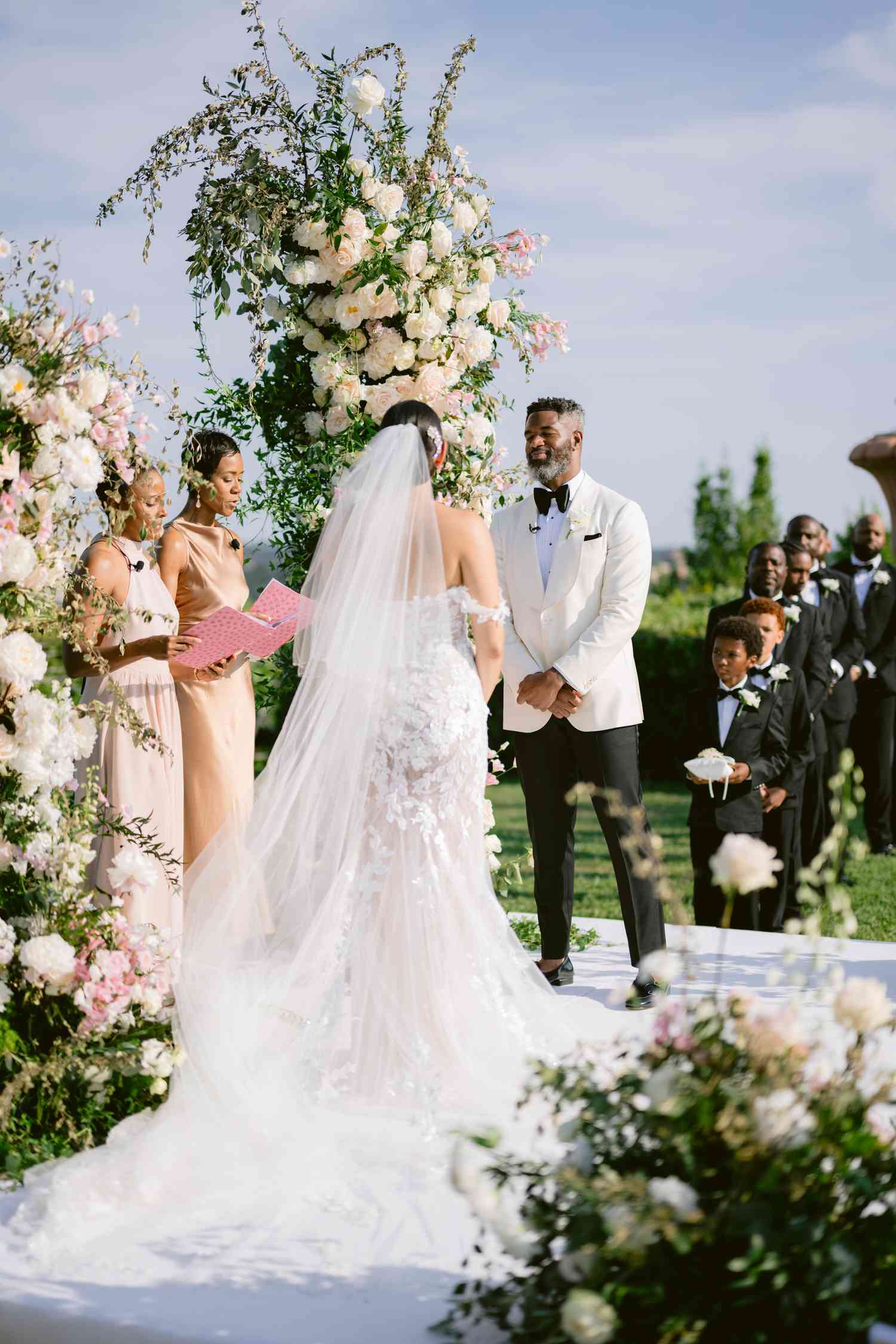 Bride in Lace Wedding Dress and Long Veil At Altar With Groom in Black and White Tuxedo Next White Flower Arches
