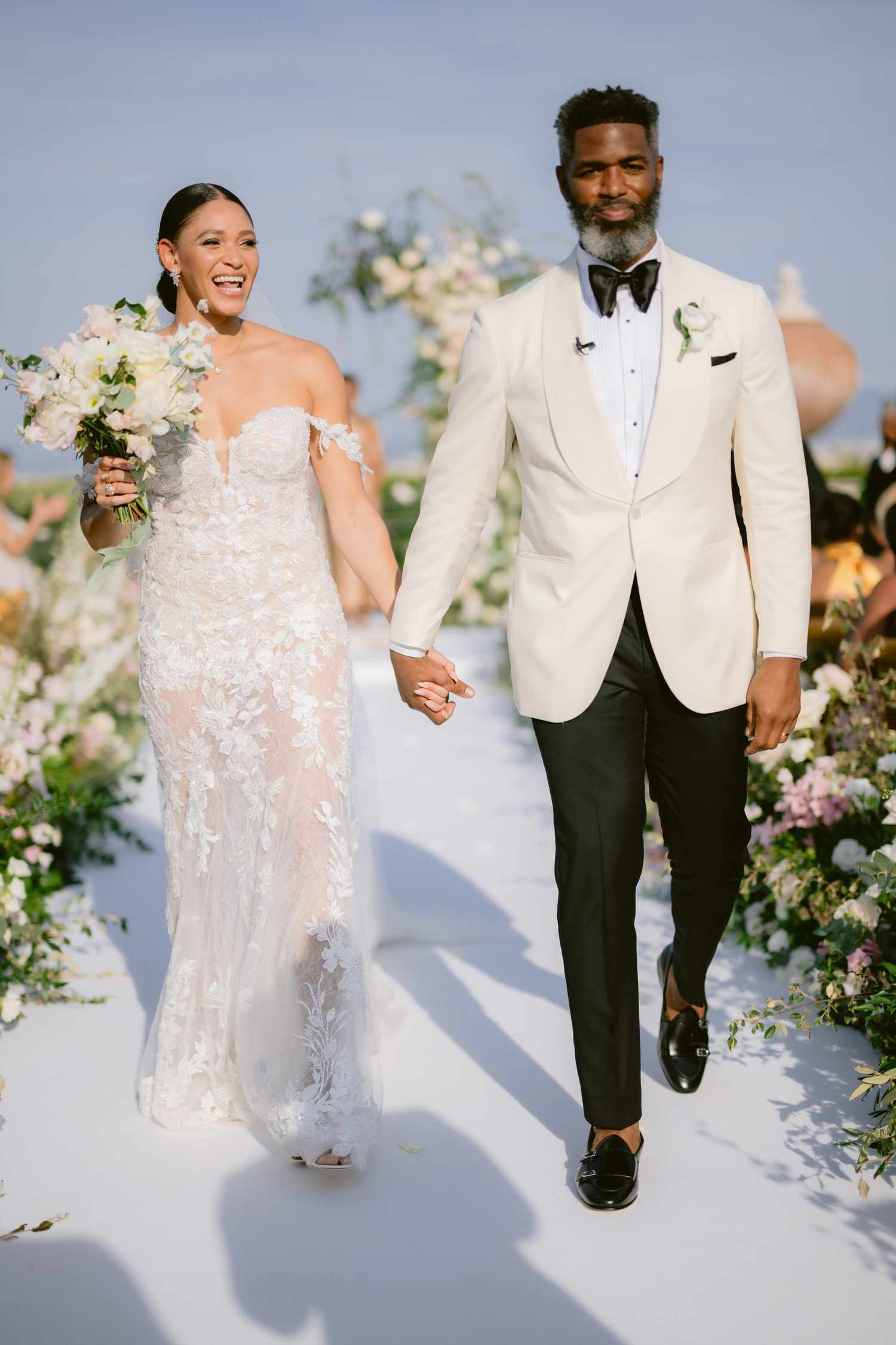 Bride in Lace Wedding Dress Holding Bouquet Holding Hands With Groom in Black and White Tuxedo
