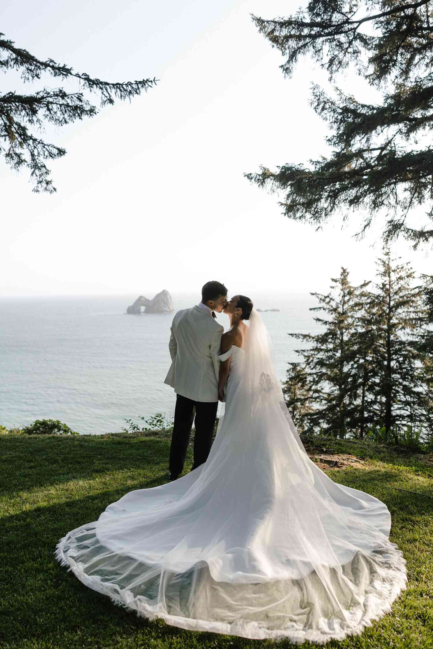 Koko lwasaki in White Off the Shoulder Wedding Dress and Veil and Kiki Nyemchek in White Tuxedo With Black Bow Tie Kissing in Front of Ocean