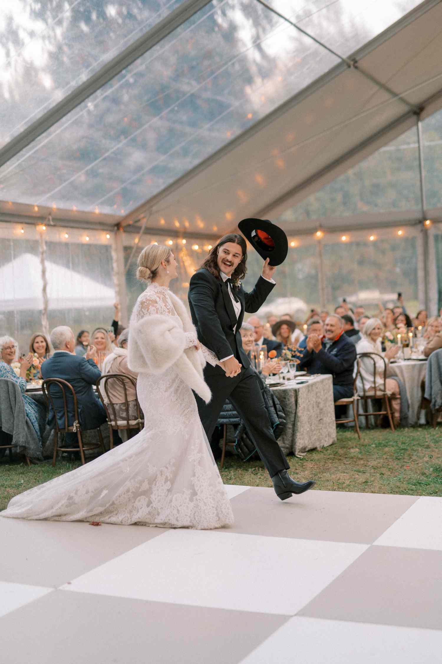 Bride in Lace Wedding Dress and Fur Coat Walking Into Reception With Groom in Black Tuxedo and Western Hat