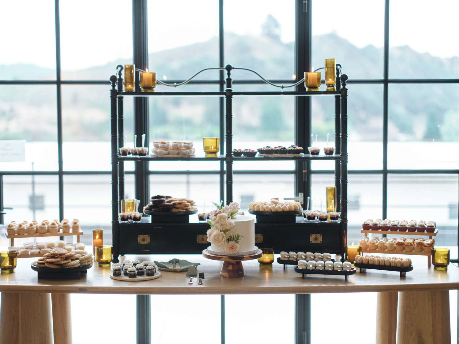 Dessert Table Spread With Small White Cake With Flowers on Top in Front of Large Glass Windows