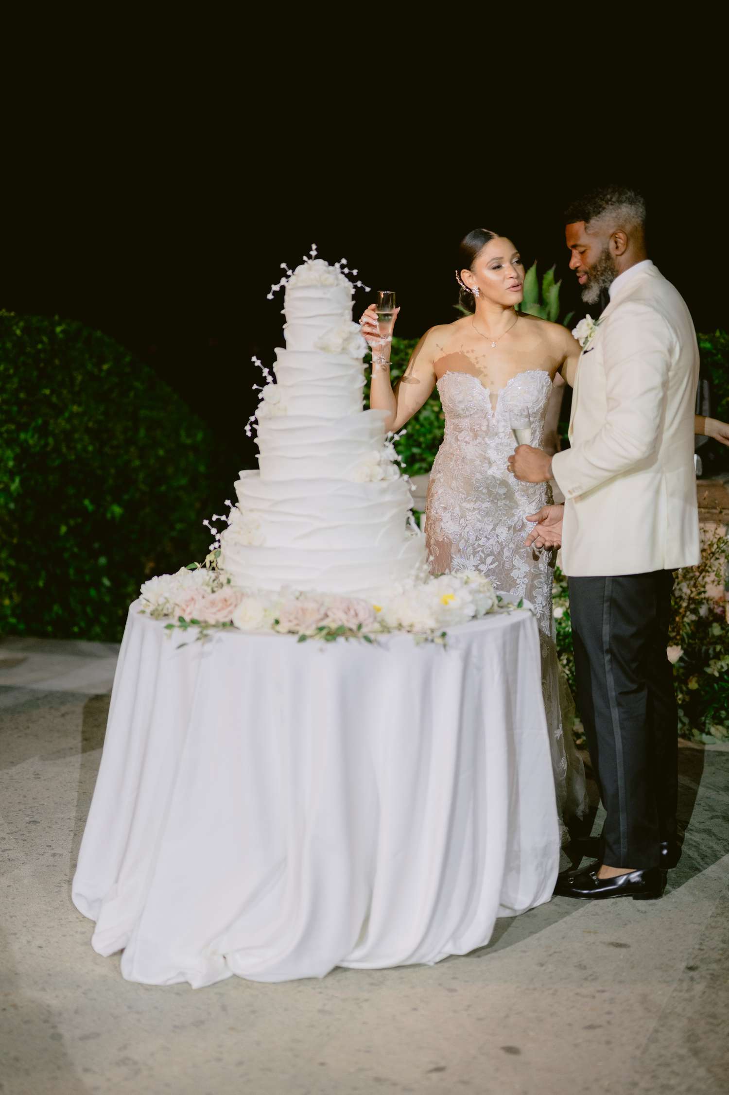 Bride in Lace Wedding Dress and Groom in Black and White Tuxedo Next to White Wedding Cake