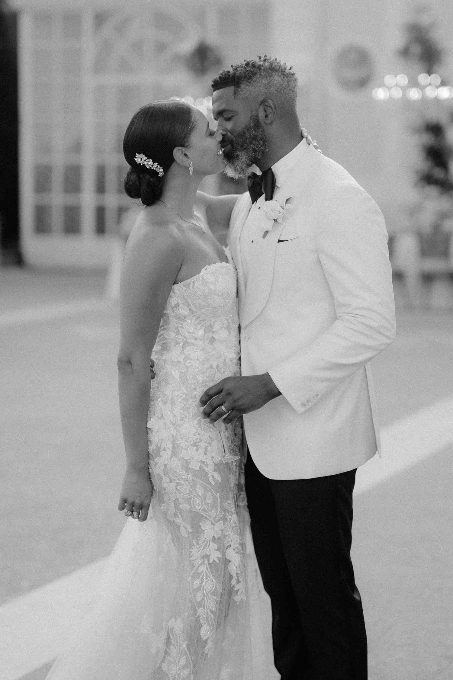 Black and White Portrait if Bride in Lace Wedding Dress and Hair in Bun Kissing Groom in Black and White Tuxedo