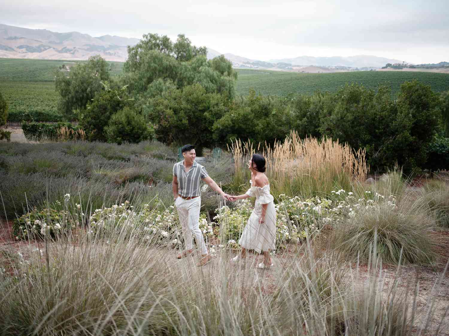 Bride in White Sundress and White Sandals Holding Hands With Groom in Stripped Shirt and White Pants in Field