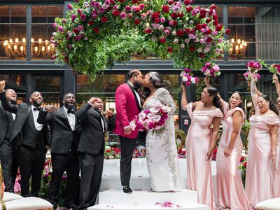 Couple kissing beneath a flower chandelier of pink and red roses with their wedding party