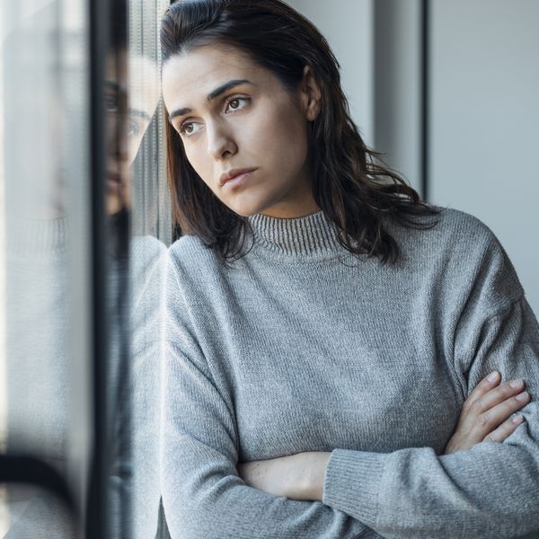 Depressed woman standing with arms crossed looking through window at home