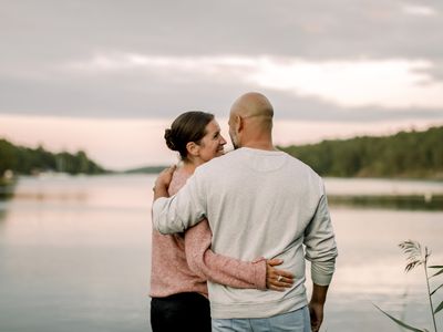 Rear view of smiling couple with arm around standing against lake during sunset